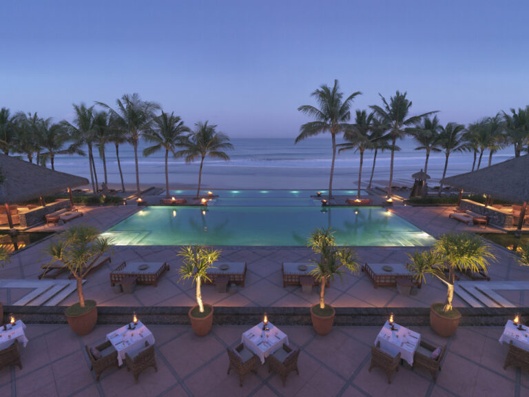 Evening view of the Legian hotel showing terrace tables set for dinner, the illuminated pools and the sea beyond.